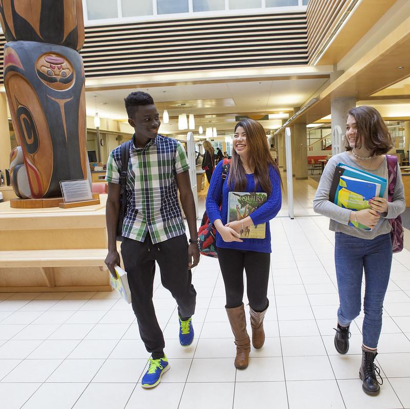 Three students walk past the totem pole in the VIU library carrying books and smiling at each other.